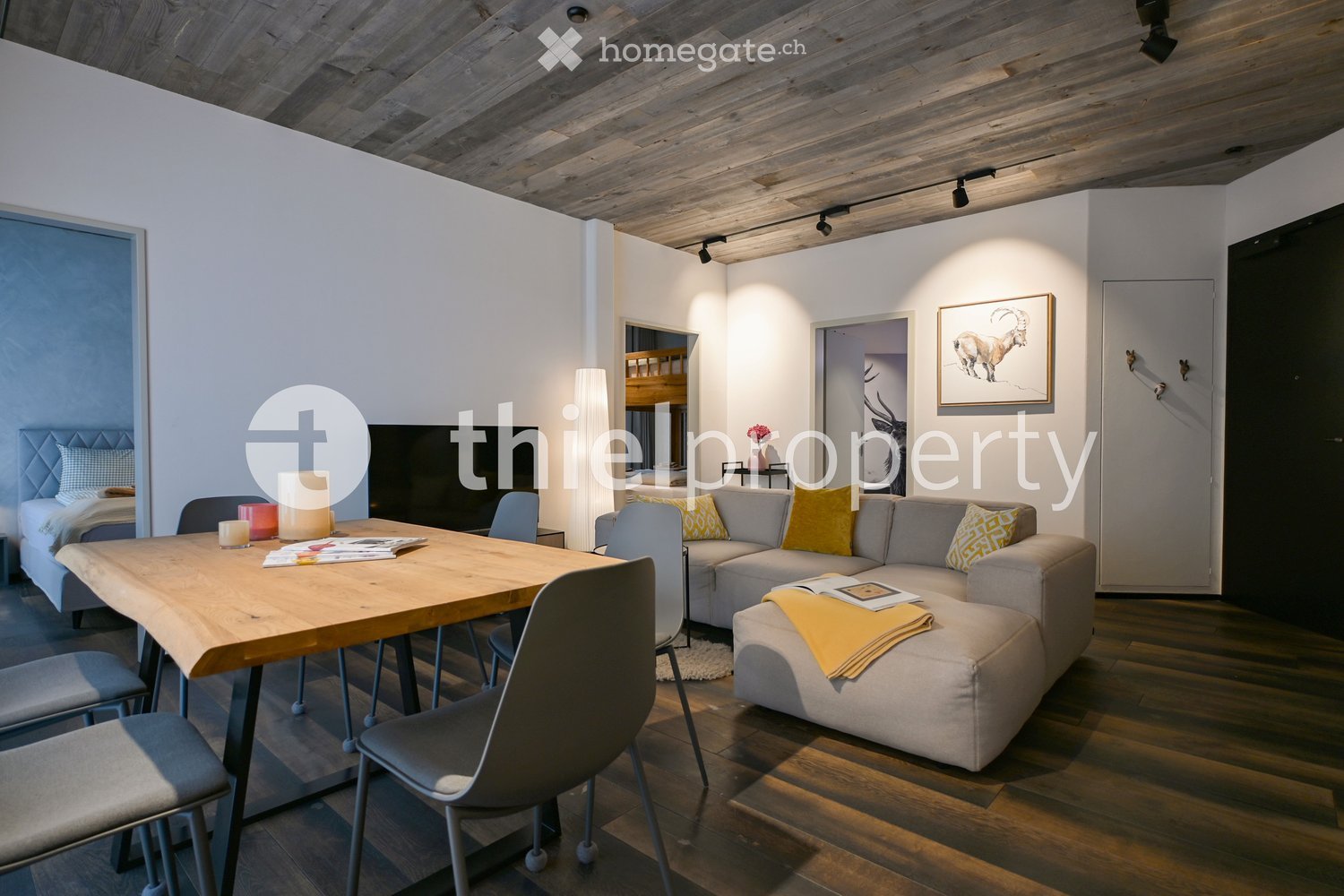 open living room with dining area, wood flooring, grey sectional sofa, dining table, lamp, and a bedroom behind the white partition