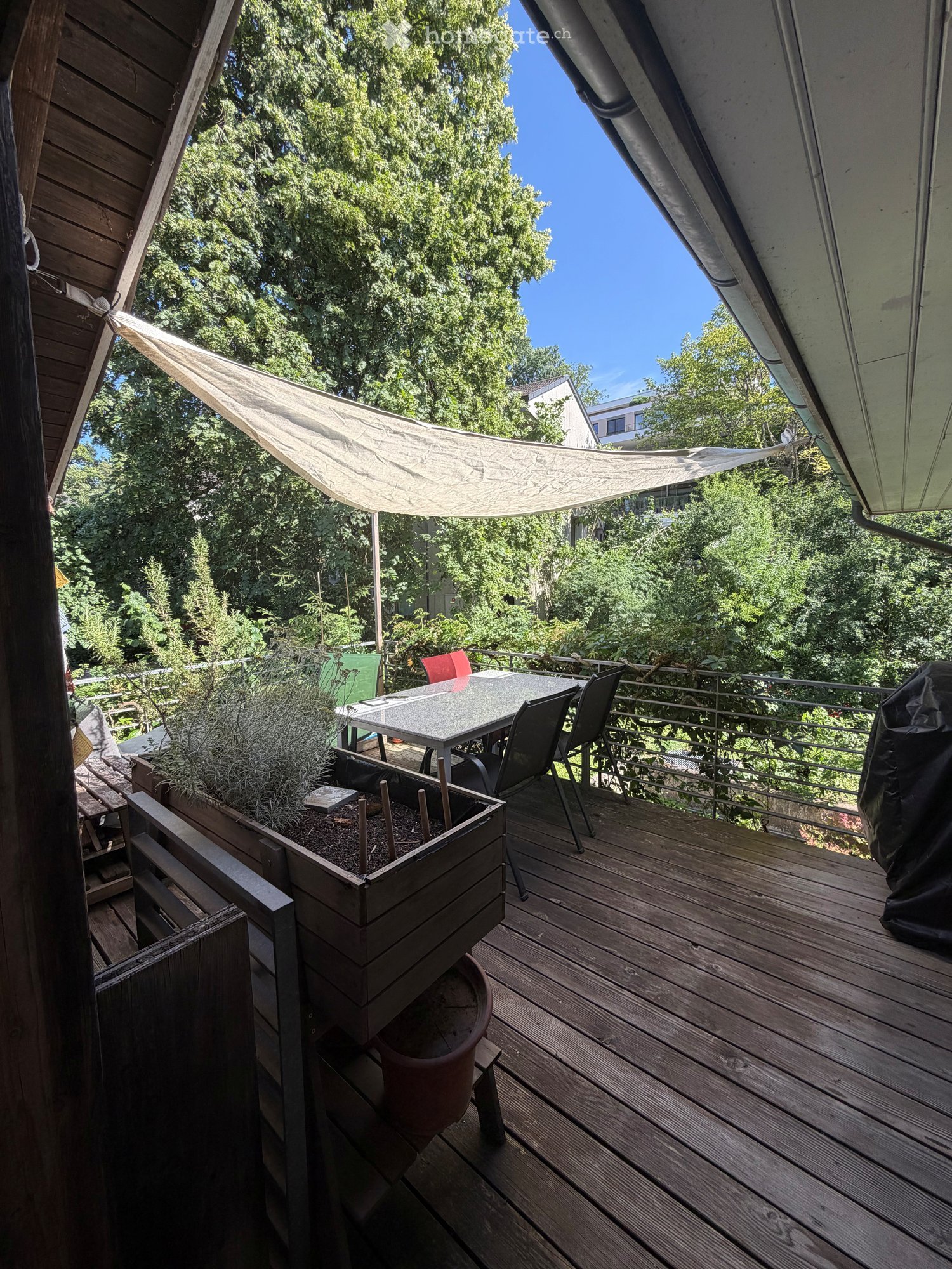 Wooden balcony with a table, chairs, and a planter, featuring a sunshade and a view of greenery.