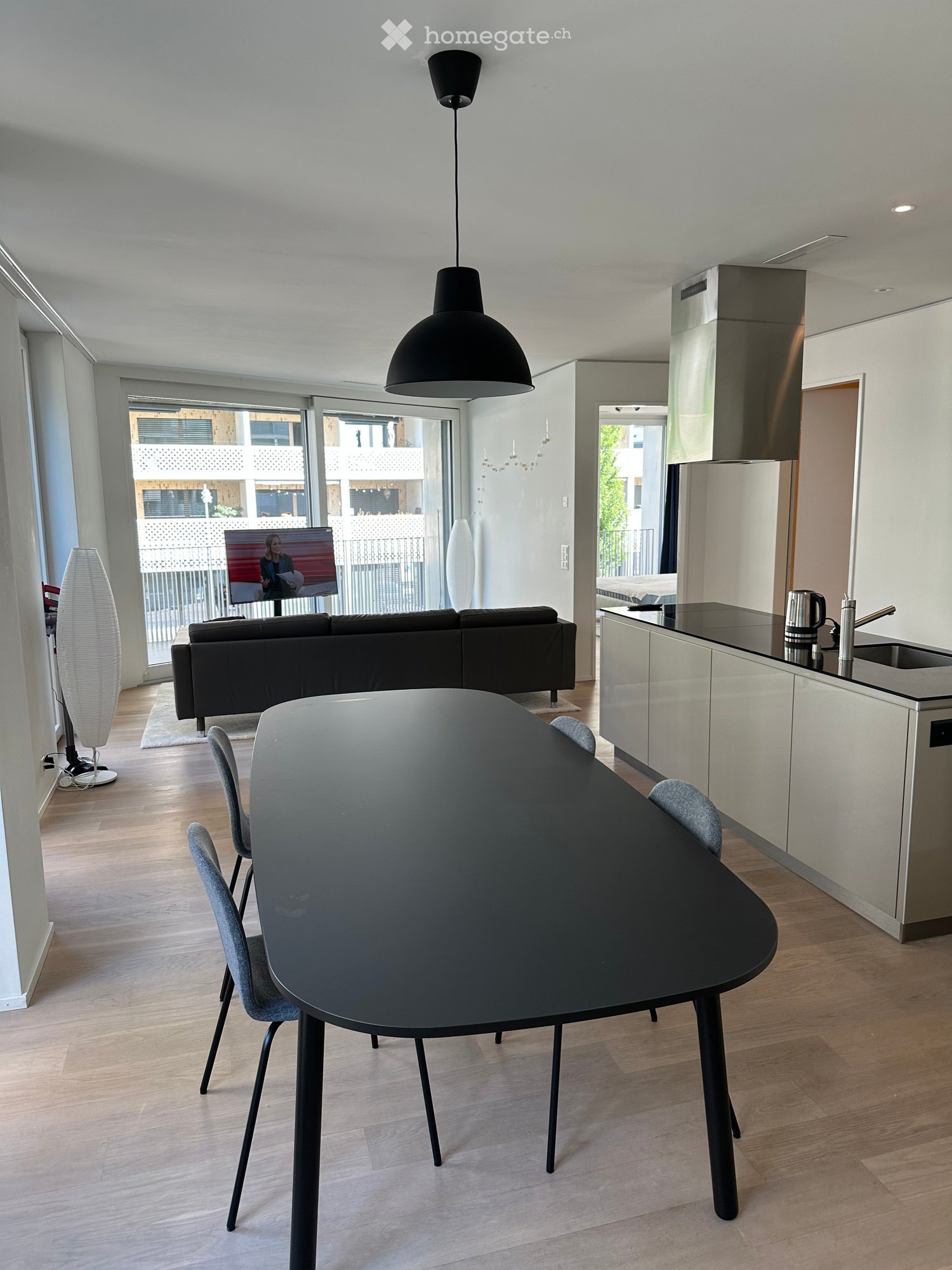 Modern living and dining area with a black sofa, a round black dining table with four chairs, a sleek kitchen area with a counter, sink, and refrigerator, and a large screen mounted on the wall. The space features light wooden flooring and large windows w