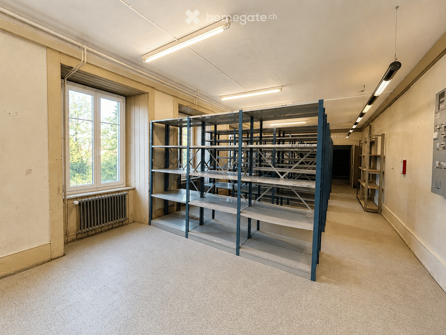 Empty room with metal shelving units, a window with a radiator below, and exposed pipes and lighting fixtures on the ceiling.
