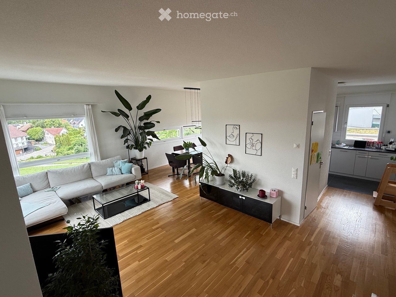 Bright living room with wooden flooring, large windows, a white sofa, coffee table, dining area with chairs and table, and a cabinet with decorative items.