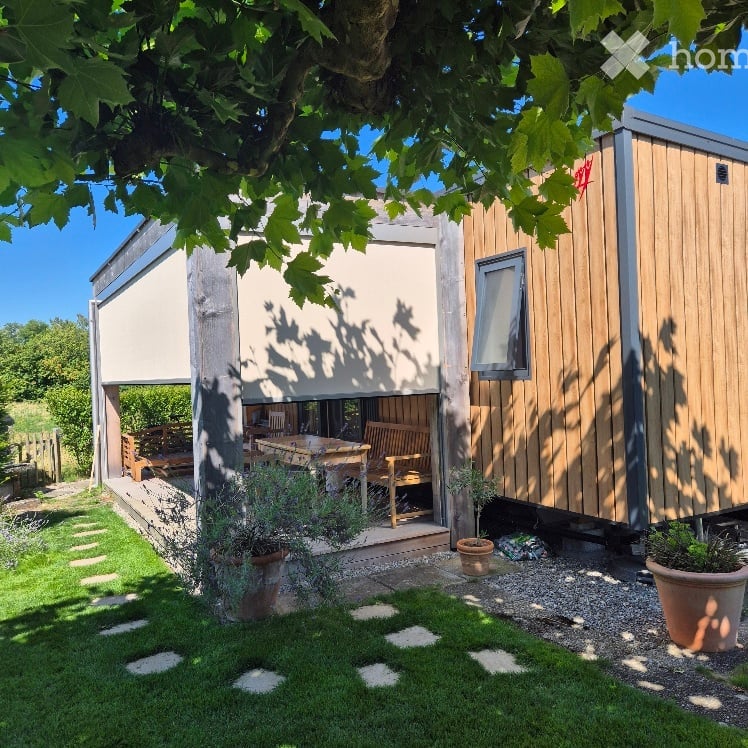 A wooden cabin with a covered patio area, outdoor furniture, and potted plants in a garden setting.