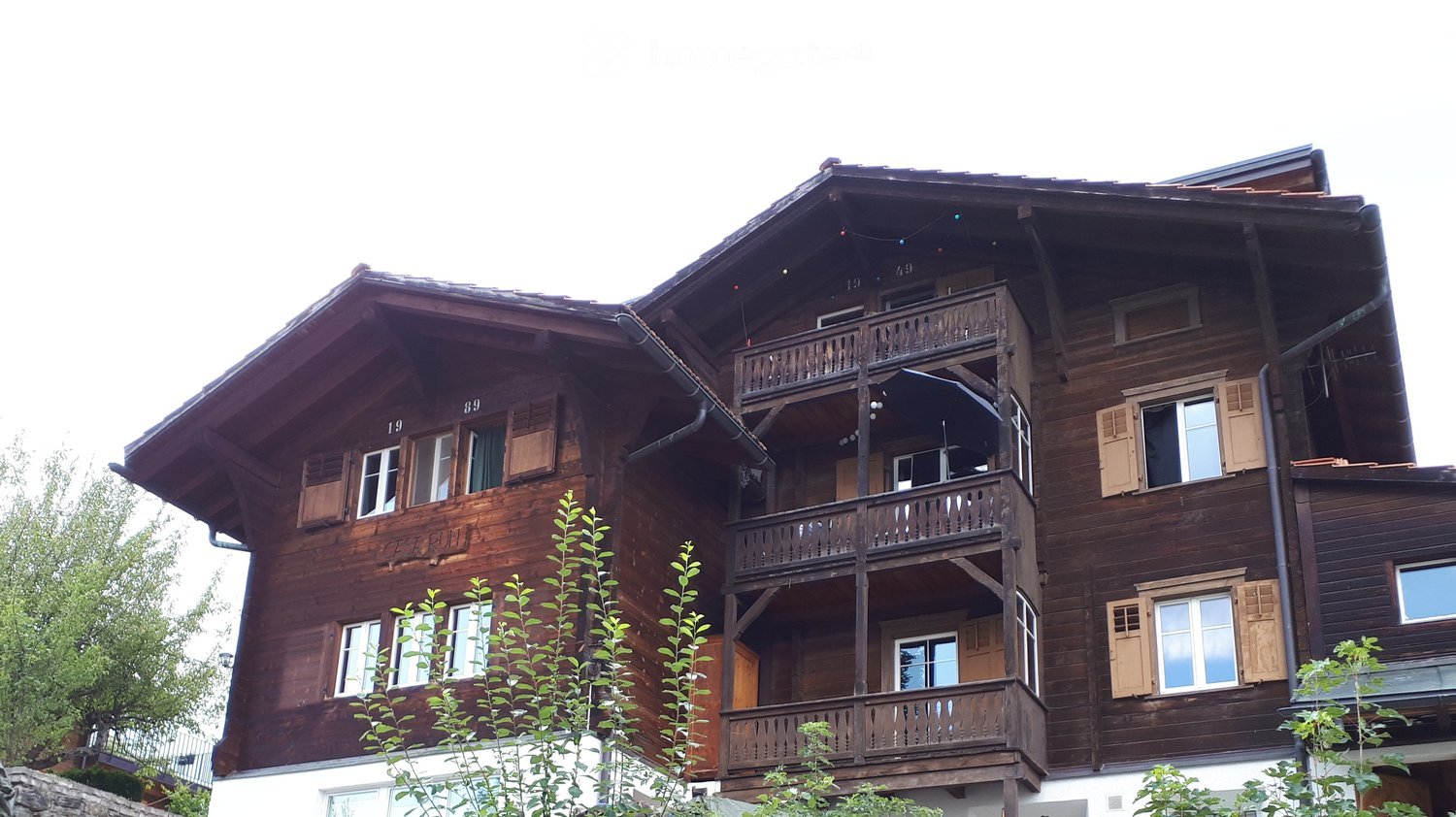 A large, multi-story wooden house with a steep, dark roof and multiple balconies. The building features numerous windows with shutters and is surrounded by greenery.