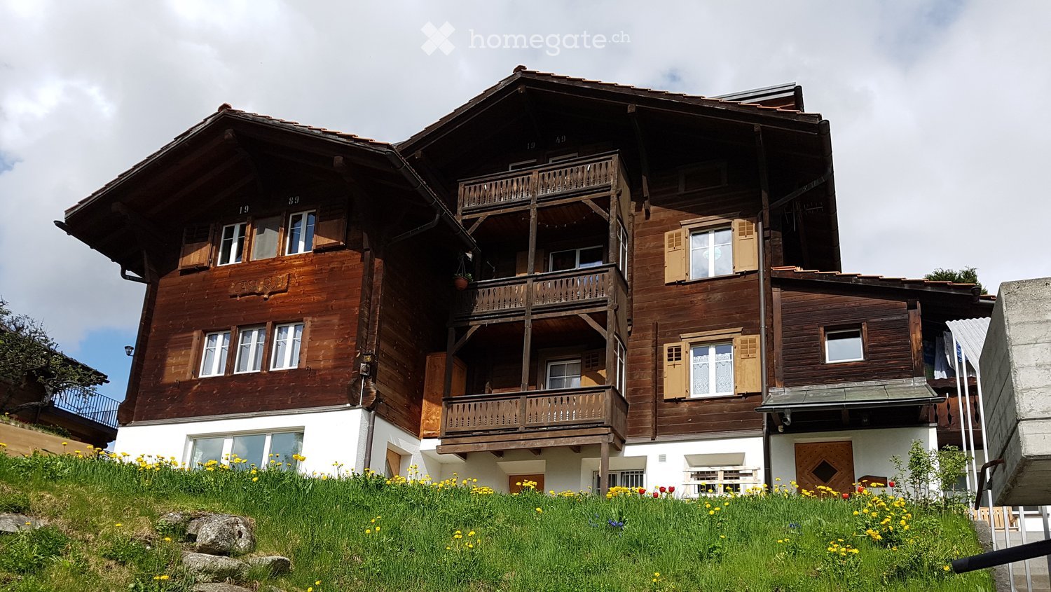 Traditional wooden house with multiple stories, balconies, and shutters, surrounded by a grassy area with yellow flowers.
