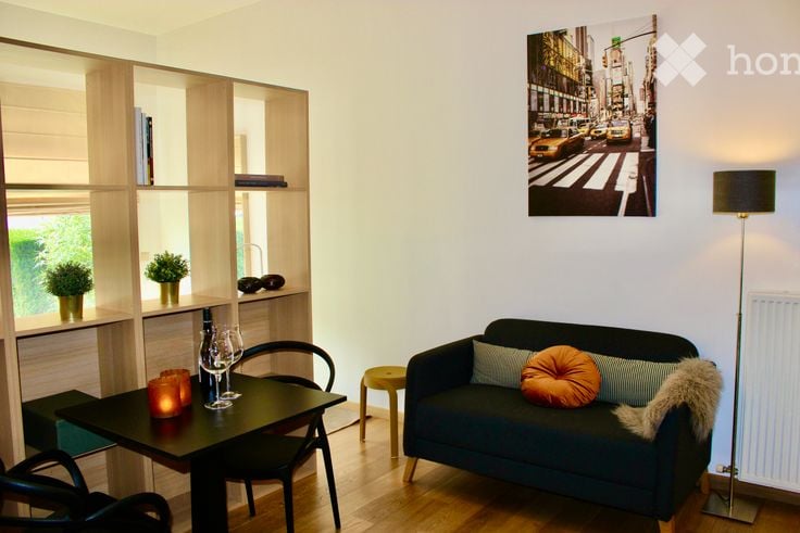 A modern, well-lit living room with a dark blue sofa, a small black dining table with chairs, a floor lamp, and built-in wooden shelves holding potted plants and decorative items.