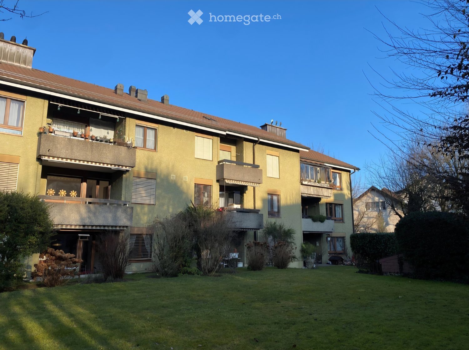 A multi-story residential building with balconies, surrounded by a well-maintained lawn and trees.
