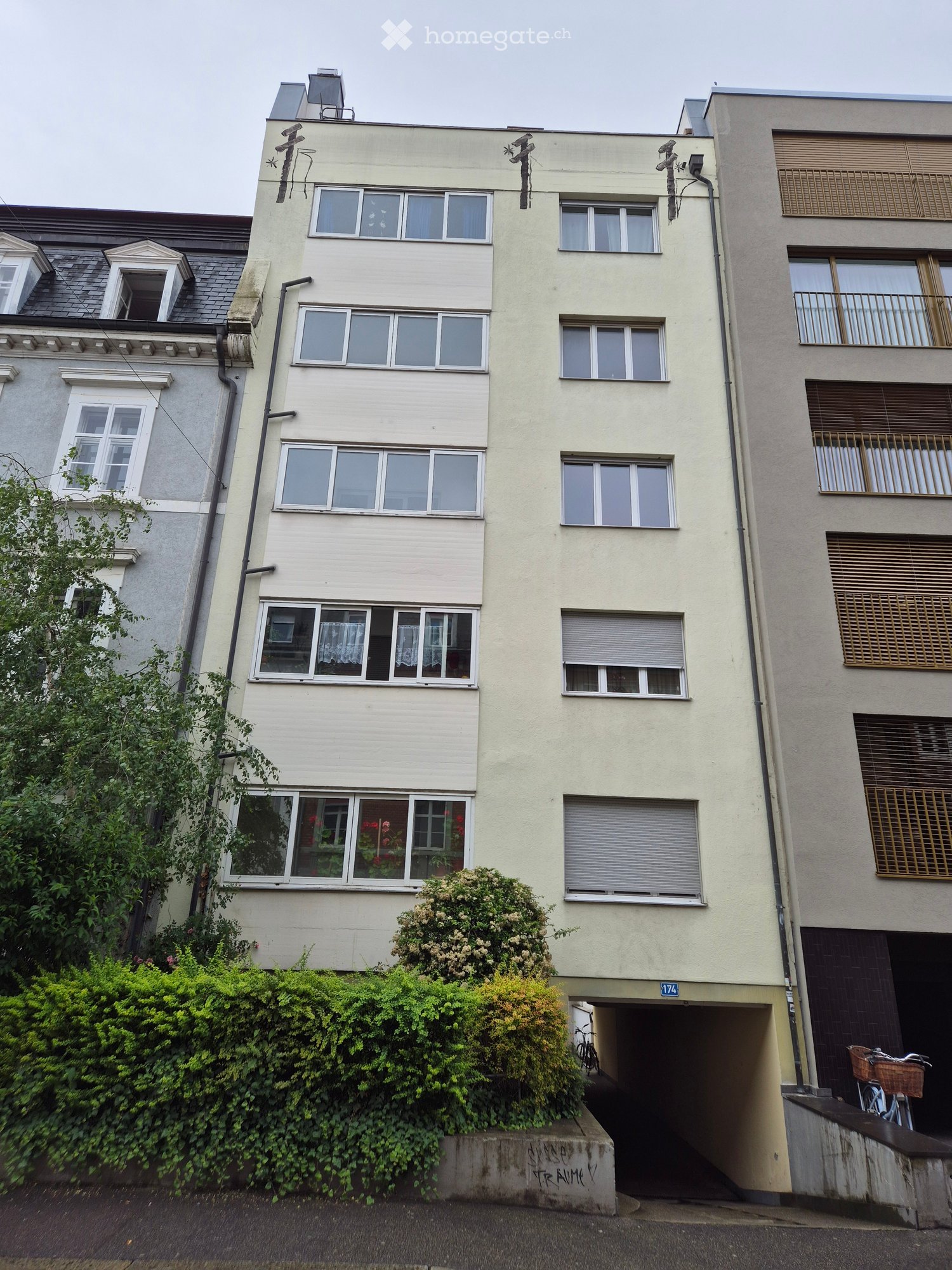 Four-story residential building with multiple windows, balconies, and a garage entrance. The facade is light-colored with decorative elements and greenery in front.