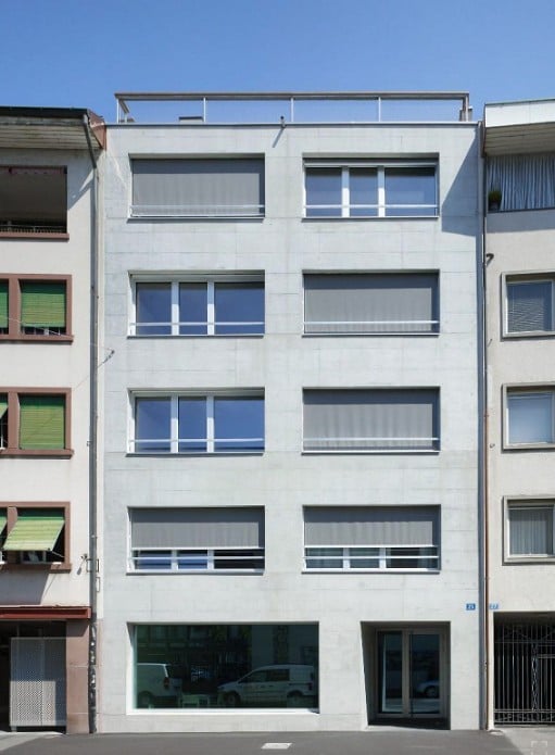 Modern multi-story building with white facade, multiple windows with blinds, and a small balcony on the top floor.