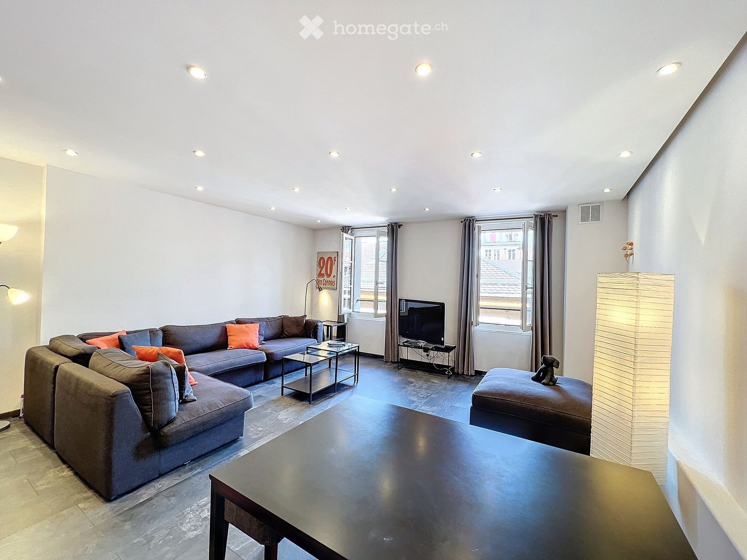 Modern living room with grey sectional sofa, orange throw pillows, coffee table, ottoman, and a television on a stand. The room features large windows with curtains and recessed ceiling lighting.