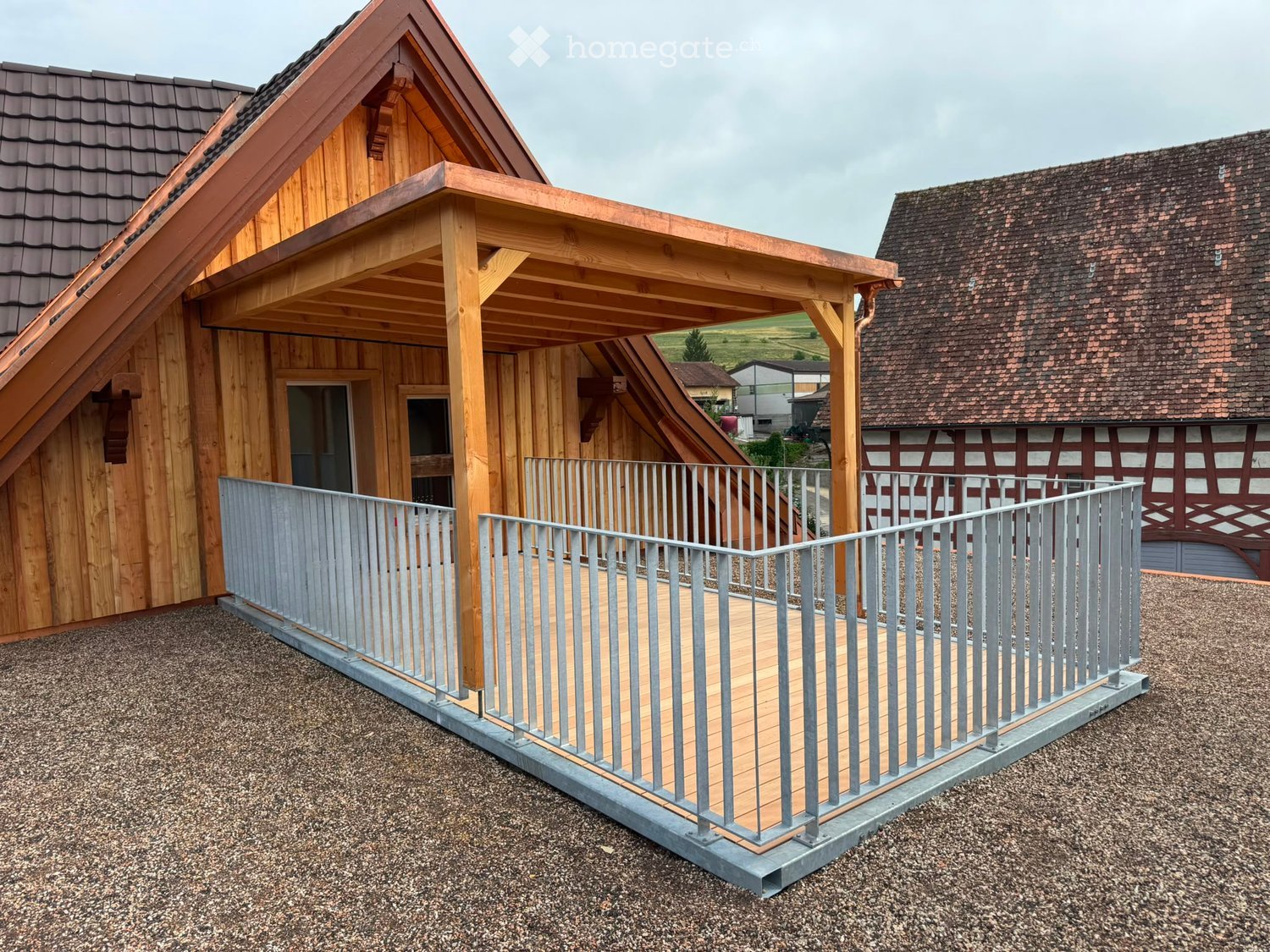 Wooden balcony with metal railing, attached to a wooden house with a sloped roof and traditional half-timbered design in the background.