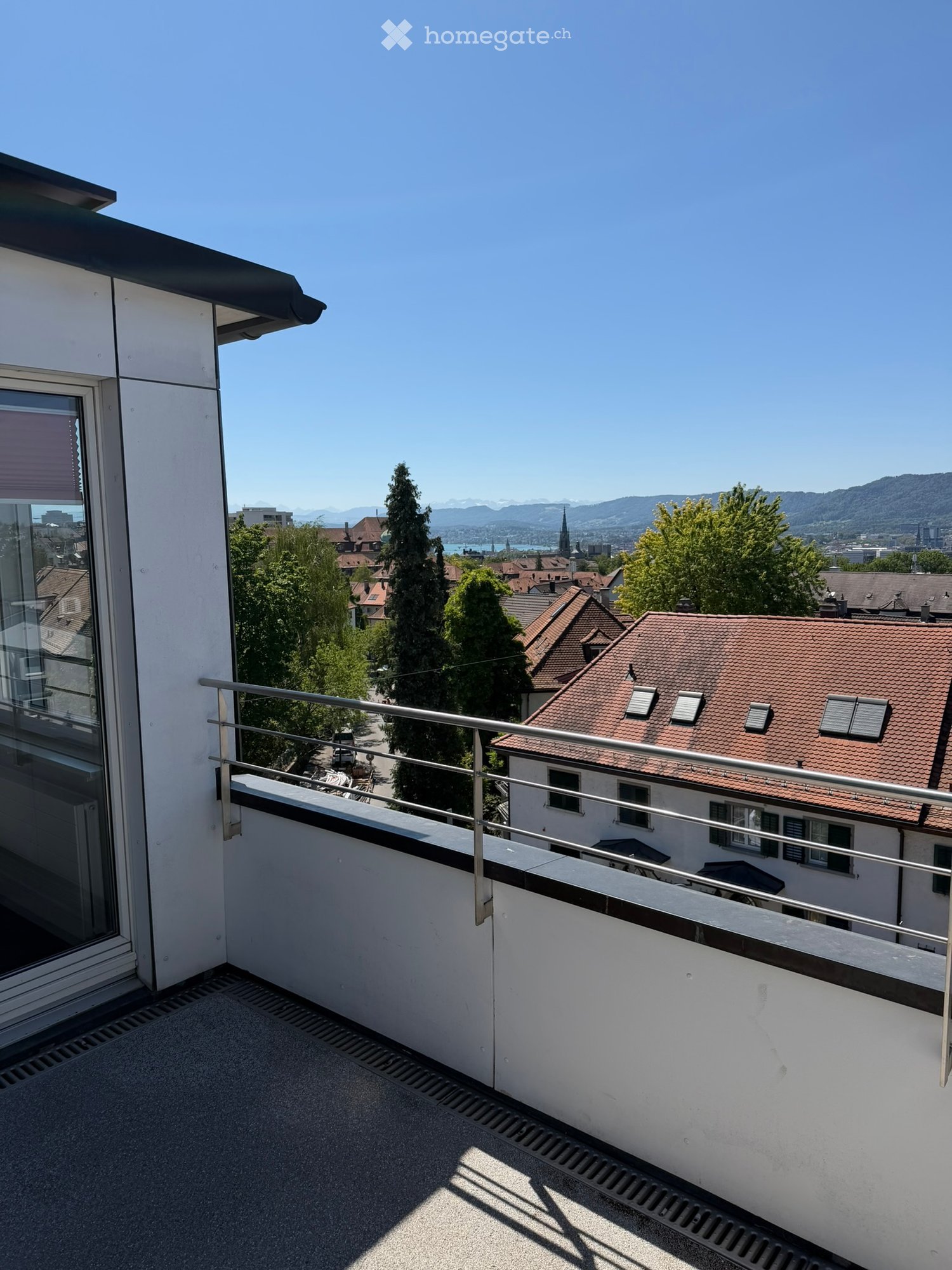 Balcony with metal railing, overlooking a cityscape with red-roofed buildings, trees, and distant mountains under a clear blue sky.