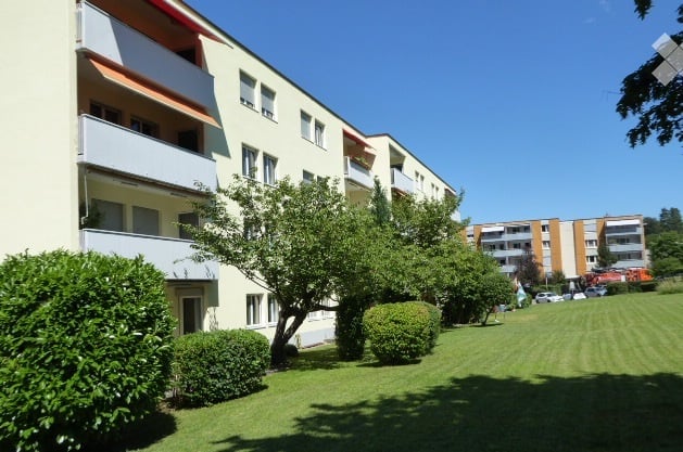 Multi-story residential building with balconies, surrounded by a well-maintained lawn and trees.