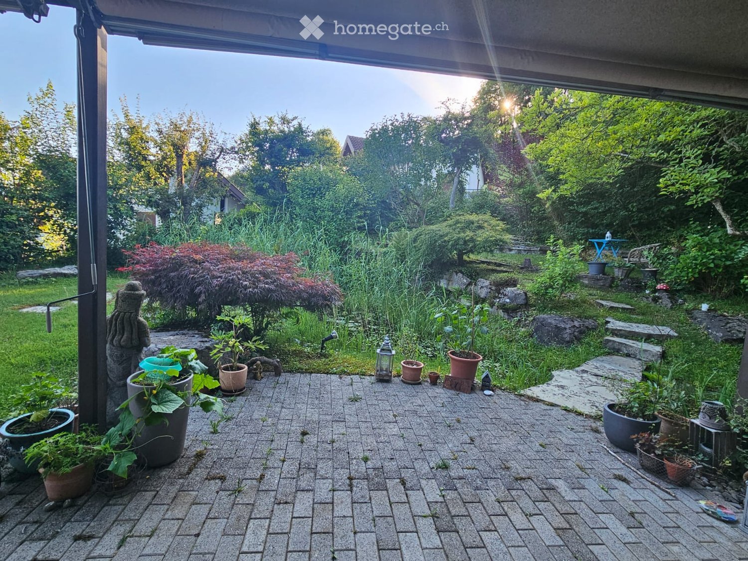 A well-maintained garden with a paved brick patio, surrounded by lush greenery, potted plants, and decorative elements like lanterns and a small statue.