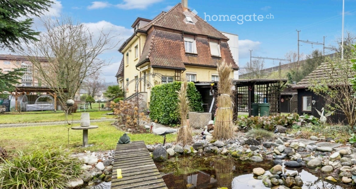 A well-maintained garden area with a small pond, decorative rocks, plants, and a wooden walkway leading to a house with a tiled roof and yellow facade.
