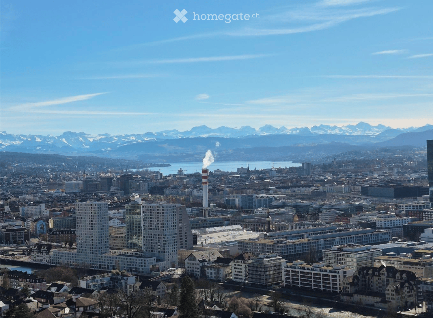 A panoramic view of a cityscape with modern high-rise buildings, traditional houses, and a distant mountain range under a clear blue sky.