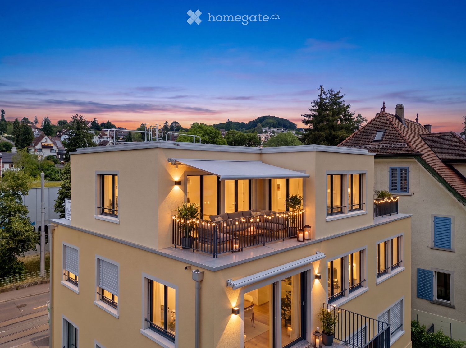 Modern multi-story building with large windows, balconies, and outdoor seating areas illuminated by string lights.