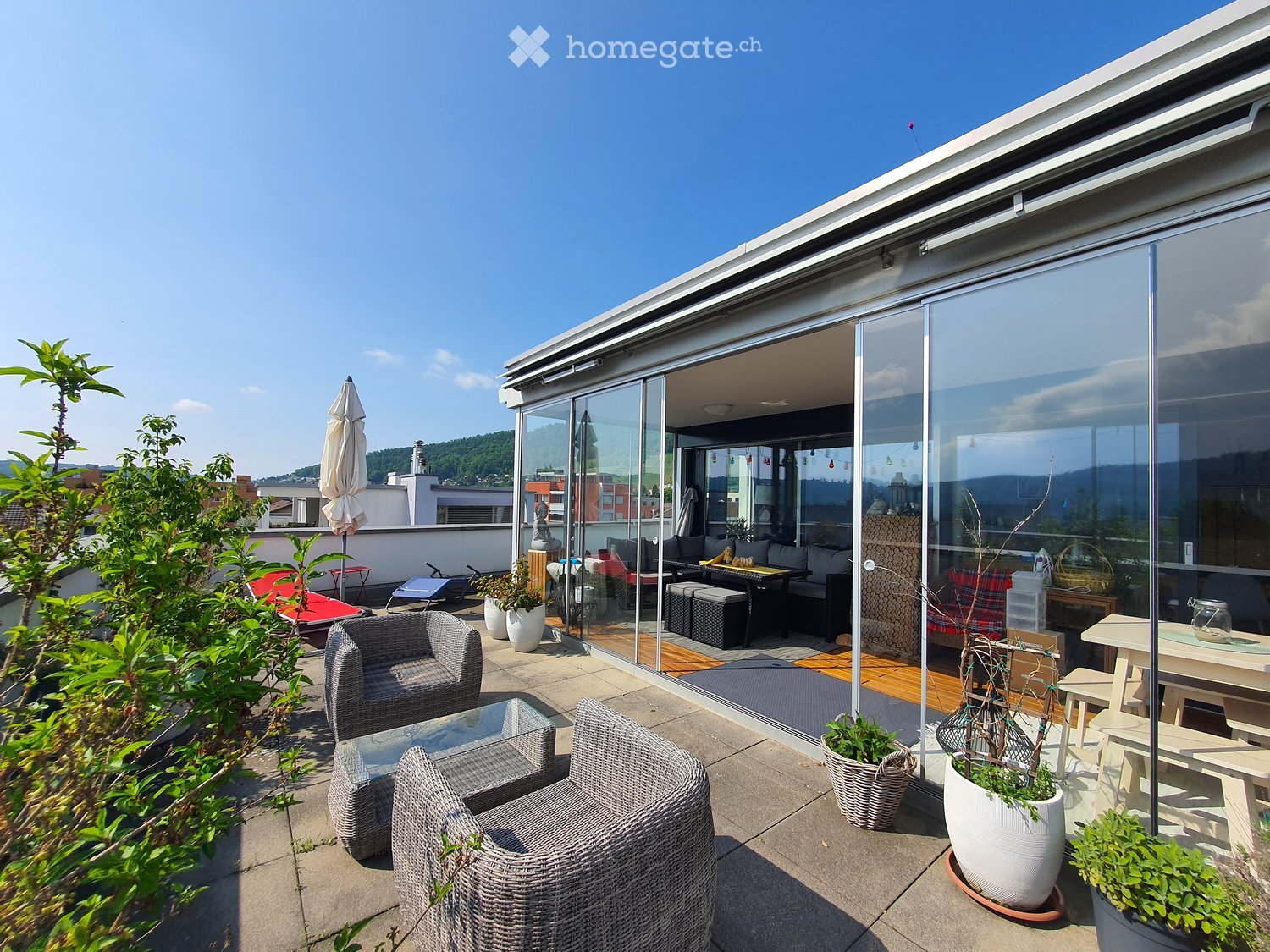 A modern rooftop terrace with wicker chairs, a glass coffee table, an umbrella, a dining table, and potted plants, connected to an indoor living space through large glass doors.