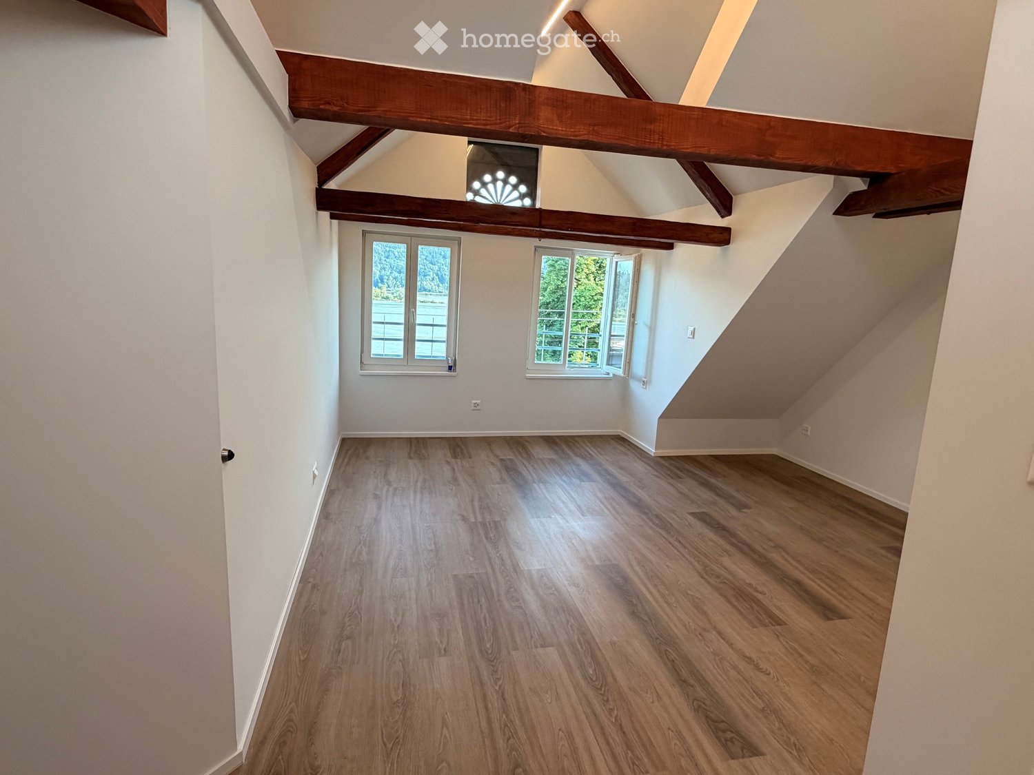 Empty attic space with wooden floors, white walls, exposed wooden beams, and two windows offering natural light.
