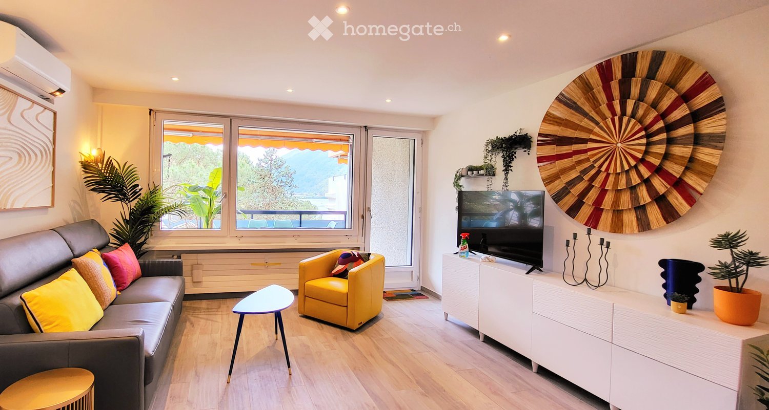 Modern living room with large windows offering a scenic view, featuring a grey sofa with colorful cushions, a yellow armchair, a small round table, a television on a white cabinet, and decorative plants.