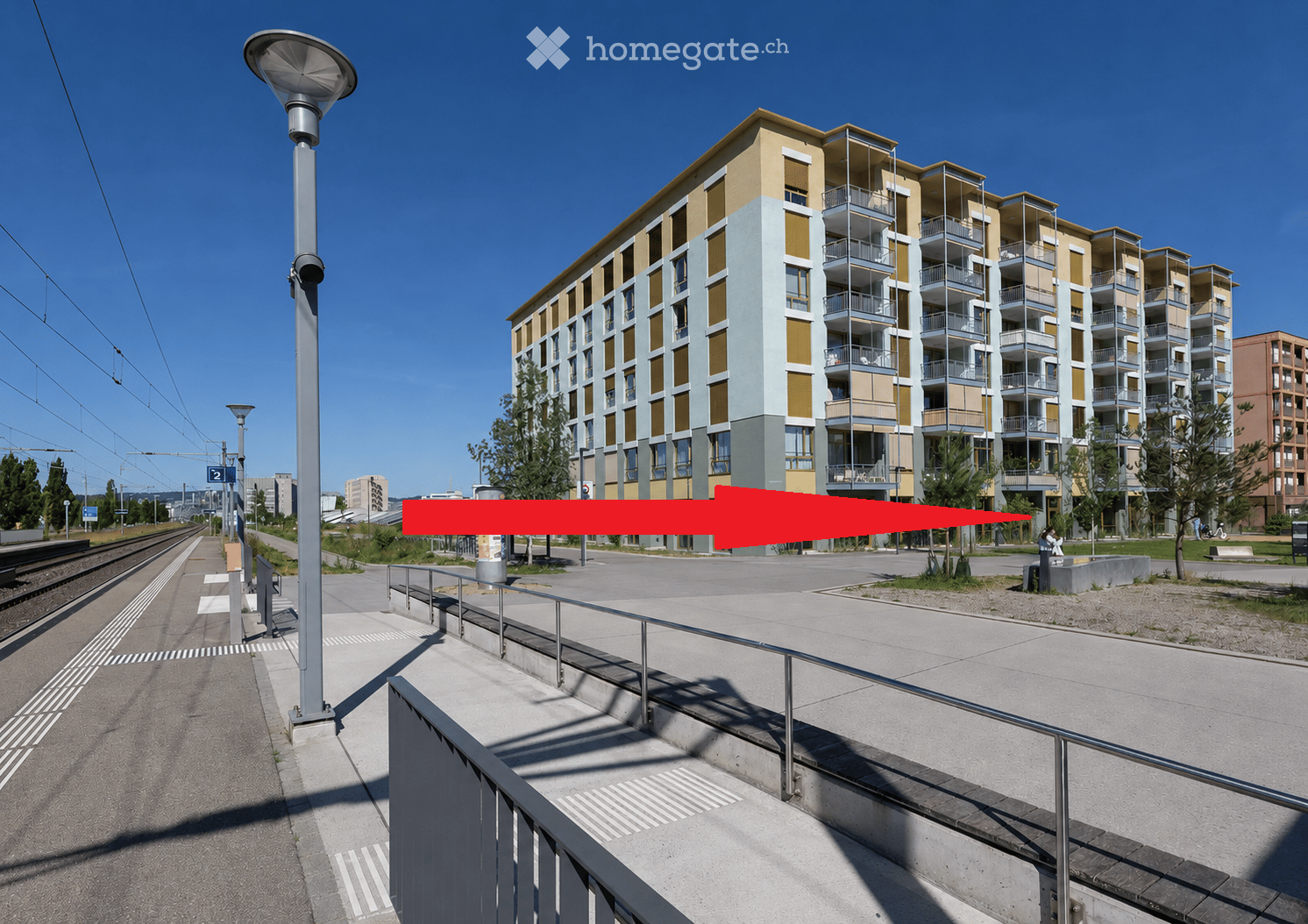 Modern multi-story residential building with balconies and large windows, located near a railway track and street with street lamps.