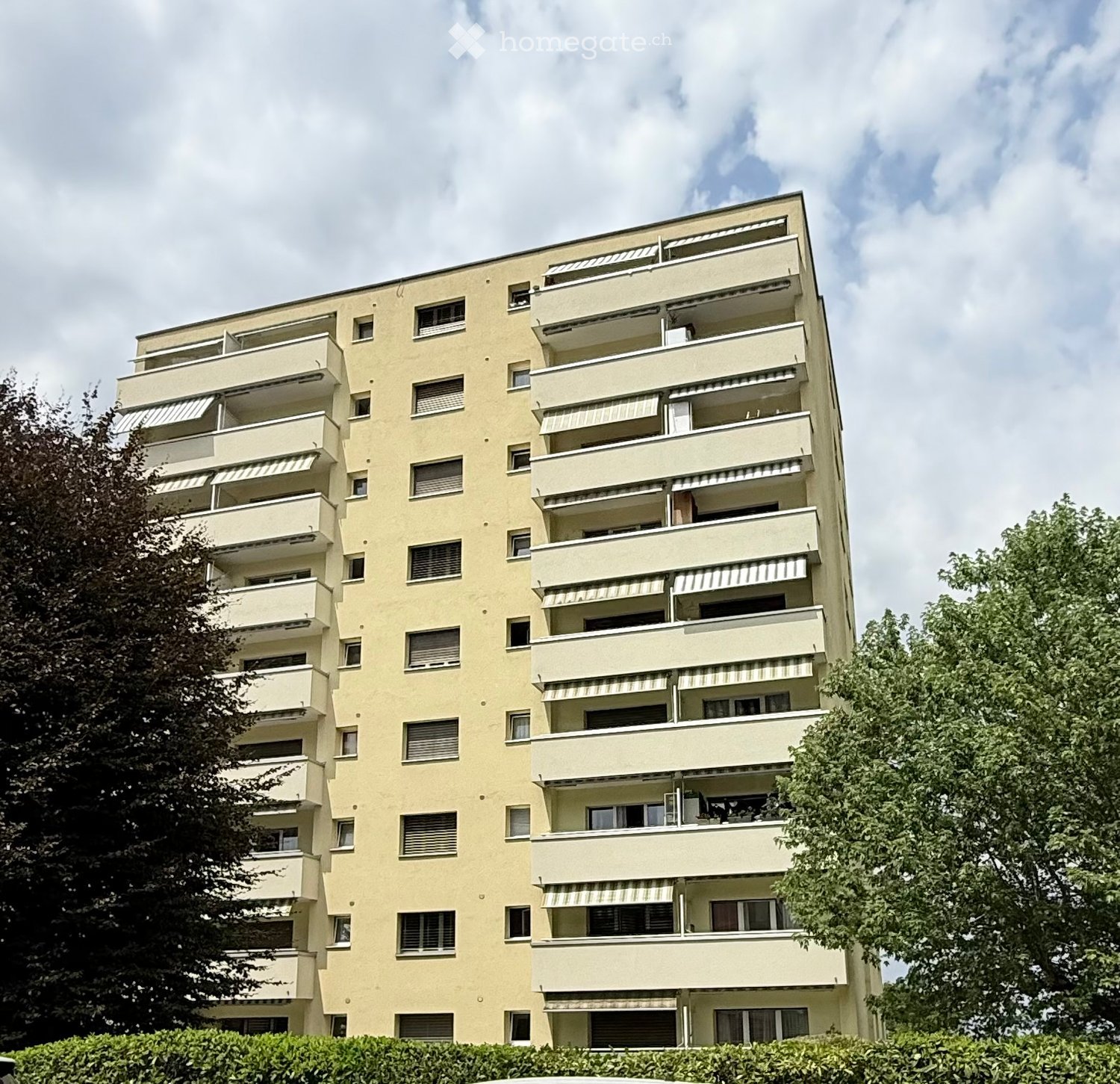 A multi-story residential building with multiple balconies and windows, surrounded by trees and greenery.