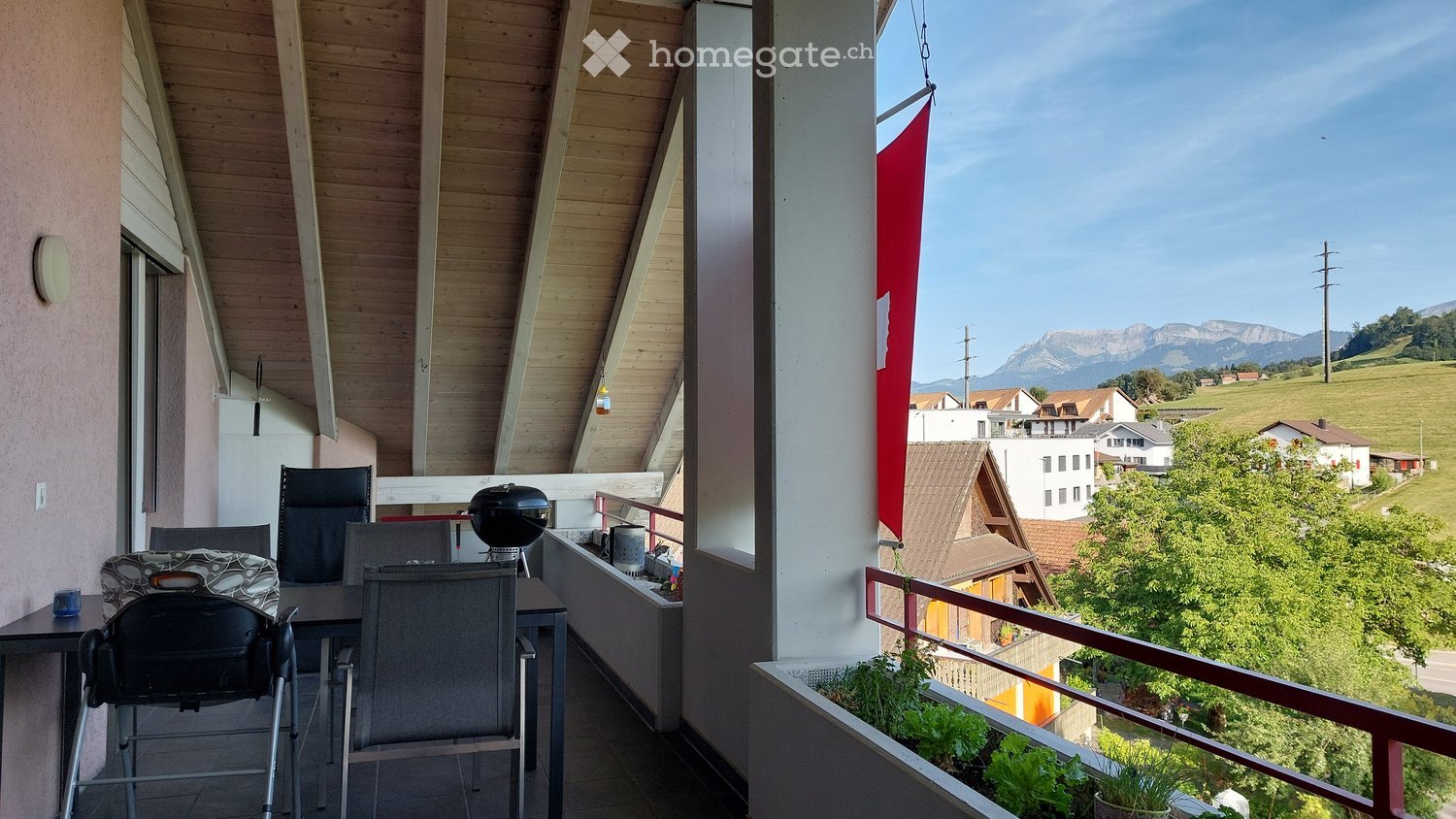 Balcony with wooden ceiling, tiled floor, outdoor furniture including a table, chairs, and a barbecue, with a scenic mountain view.