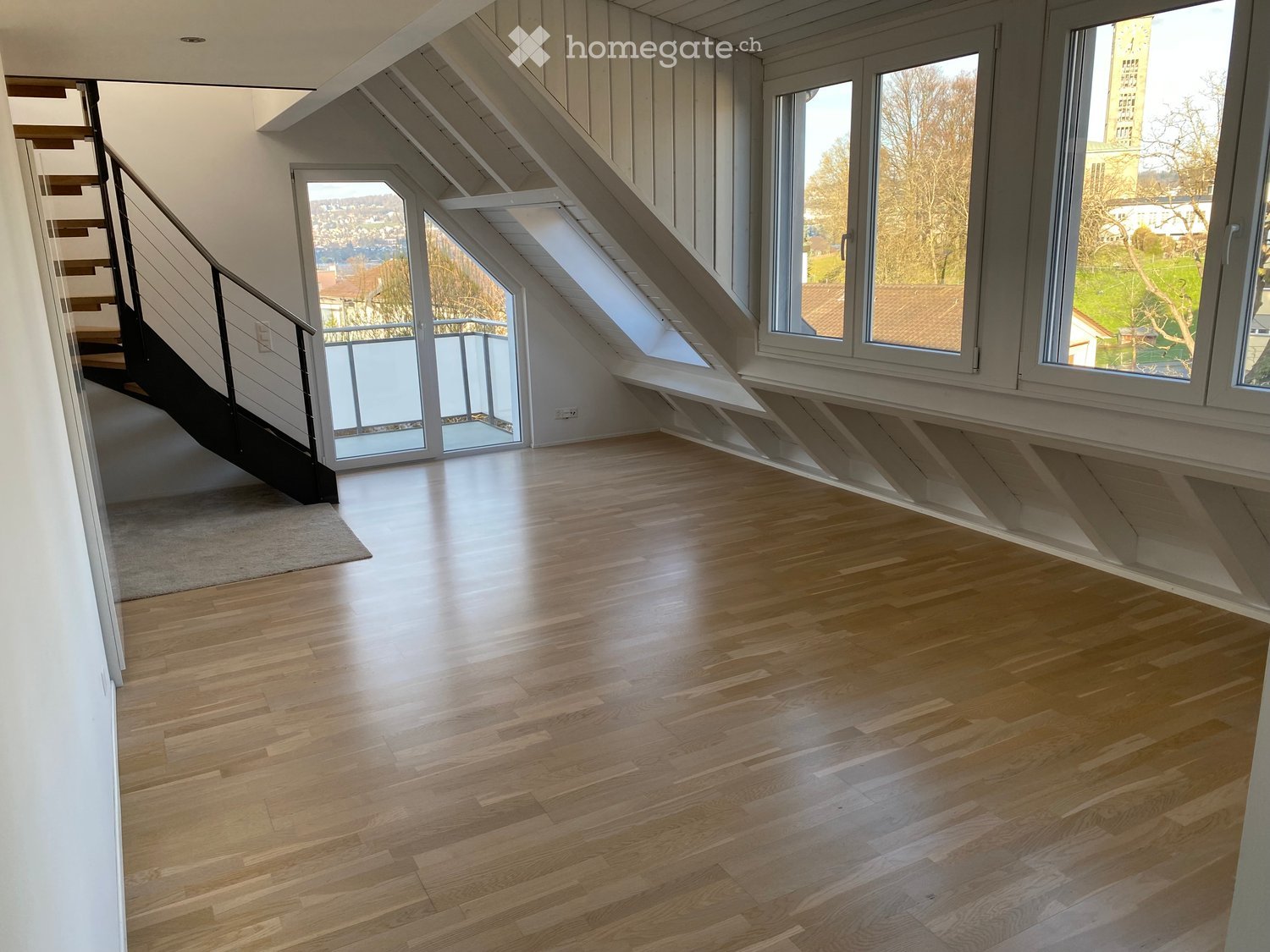 Empty room with wooden flooring, white walls, large windows, and a staircase leading upwards.