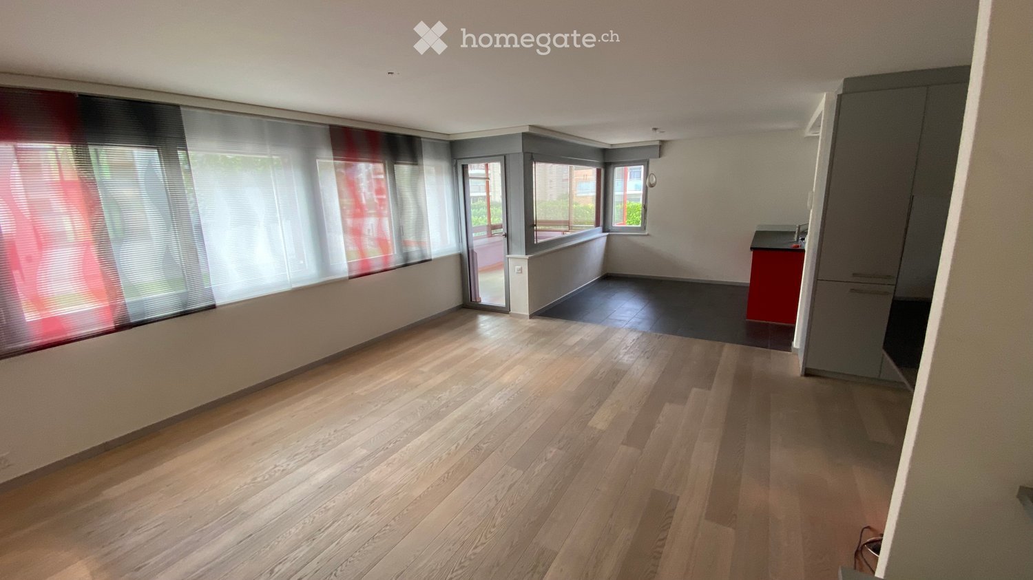 Empty room with wooden flooring, large windows with blinds, and a small kitchen area with cabinets and a countertop.