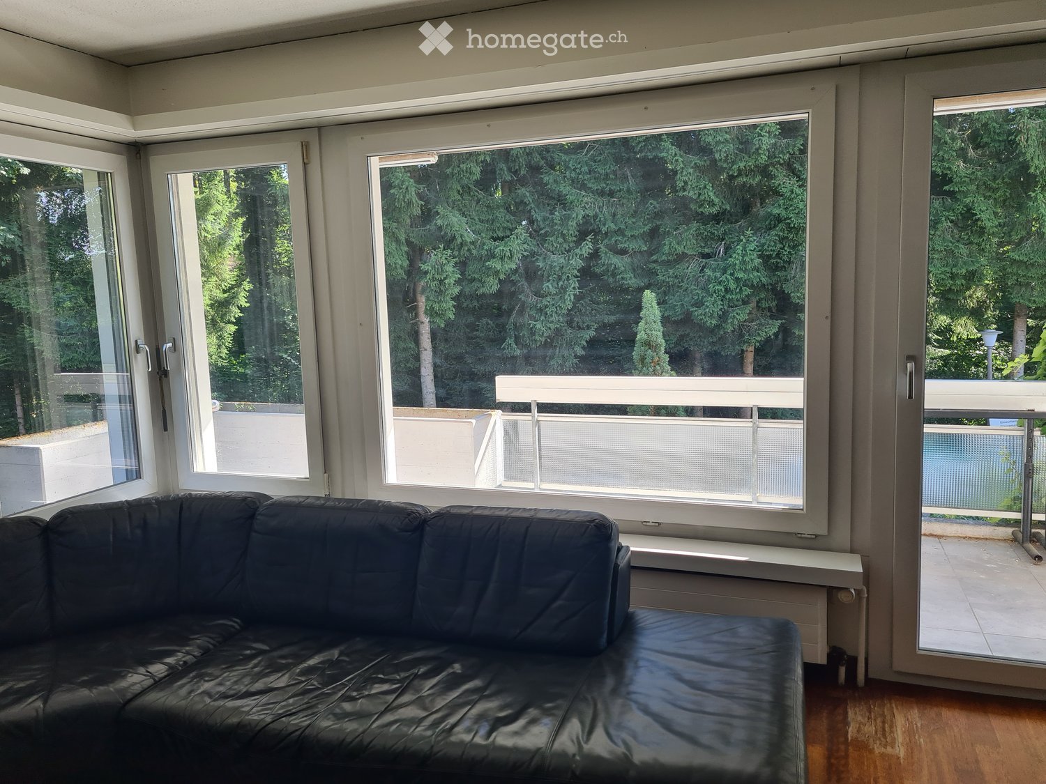 Modern living room with large windows and a balcony, featuring a black leather sofa and wooden flooring.