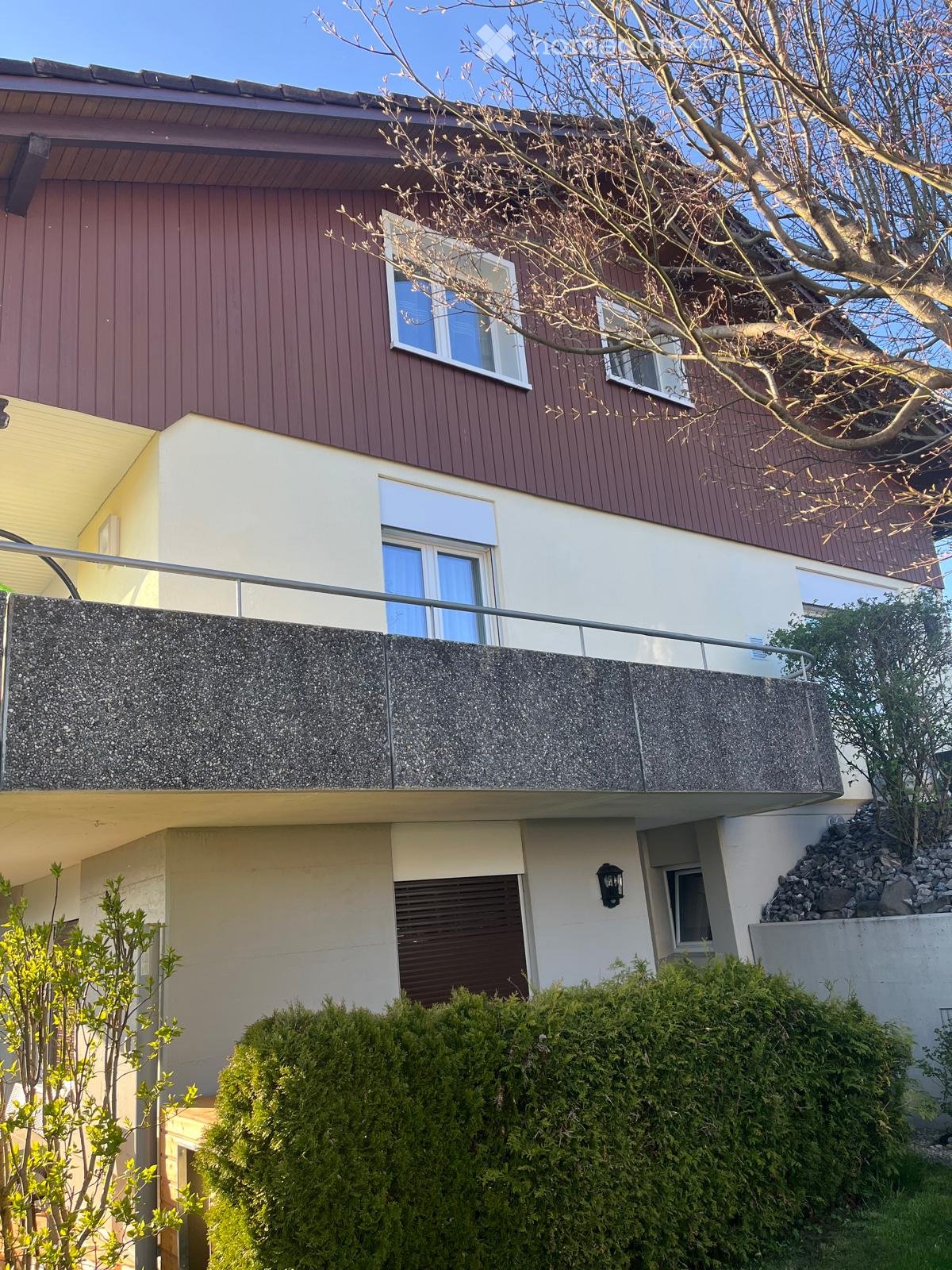 Two-story house with red-brown siding, white trim, multiple windows, and a balcony with a metal railing.