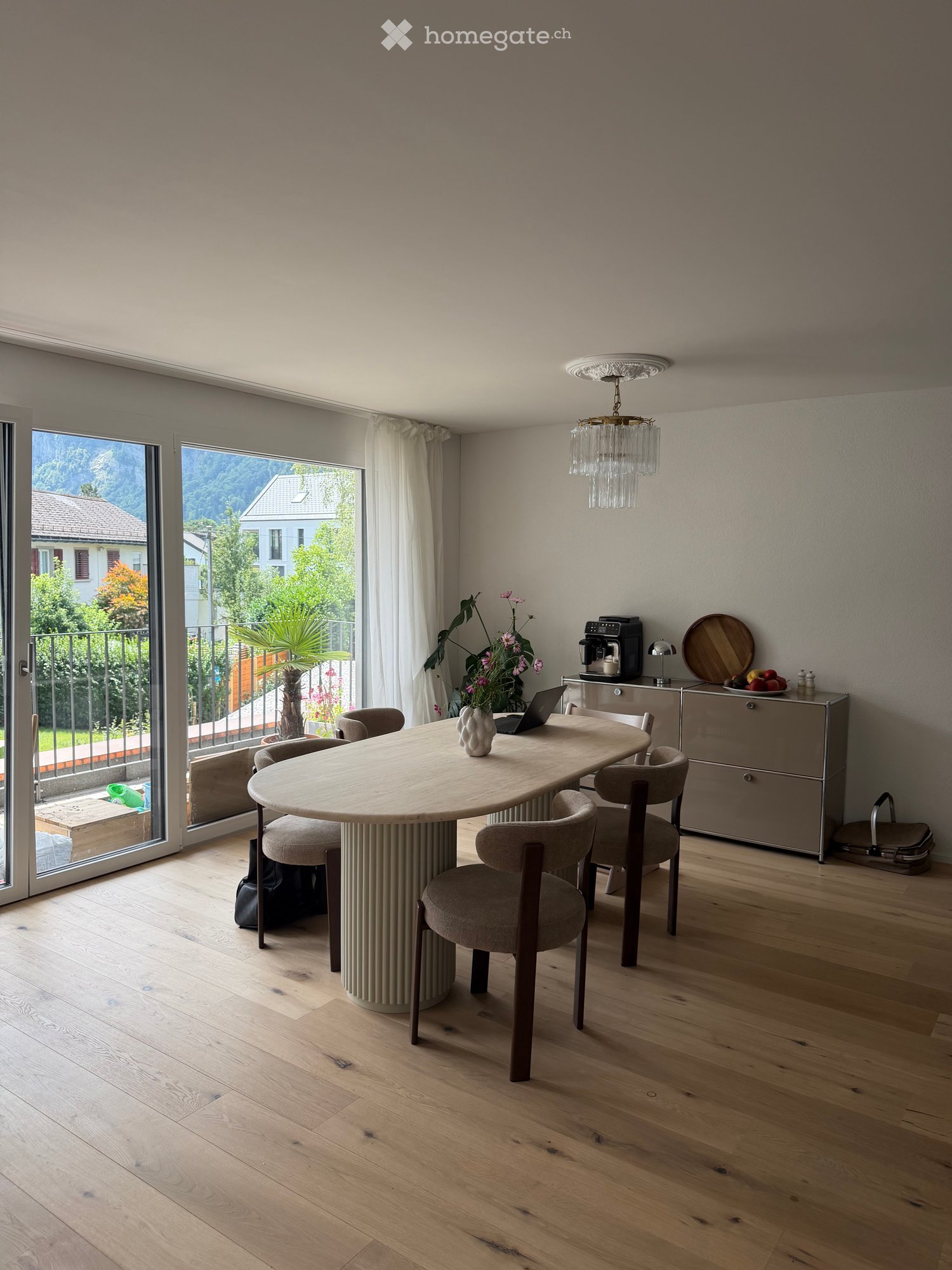 Modern kitchen with wooden floor, large round table, six chairs, laptop, vase, coffee machine, cabinet, and a view of the outdoor area.