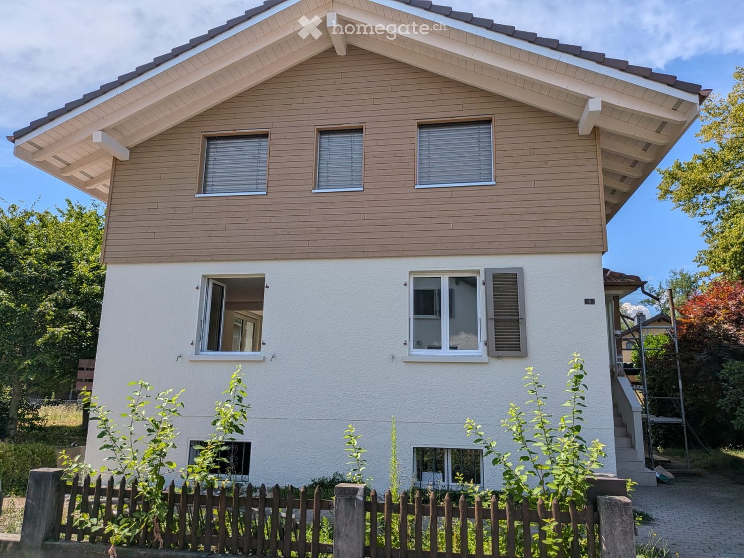Two-story house with wooden siding on the upper level and white plaster on the lower level, featuring multiple windows, a small porch, and a wooden fence in the front yard.