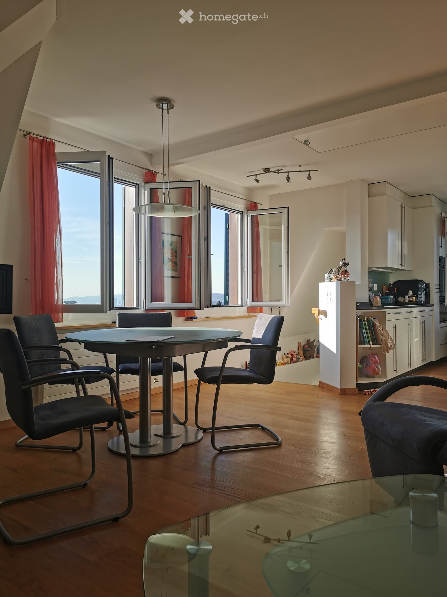 A modern, well-lit room with wooden flooring, large windows with red curtains, a round glass table with four chairs, a sofa, and a small kitchen area with cabinets and a refrigerator.