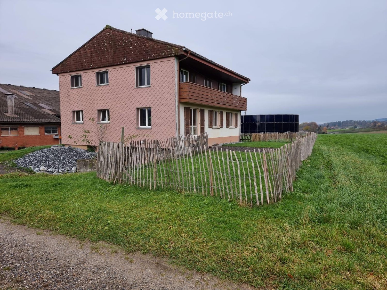 Two-story house with pink siding, multiple windows, and a wooden balcony. Surrounded by a wooden fence and grassy field.