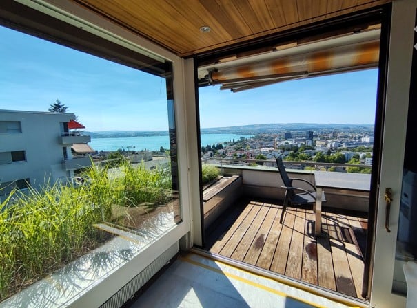 Balcony with wooden decking, large windows, scenic city view, and a single chair.