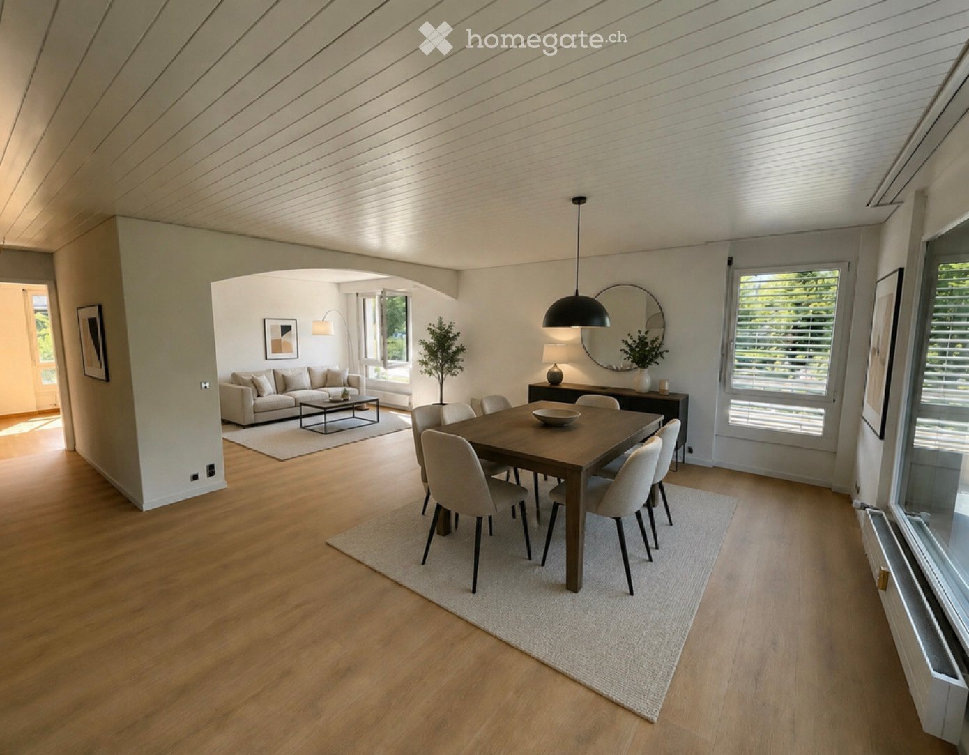 Open-concept living and dining area with light wood flooring, a beige sofa, coffee table, dining table with chairs, and modern decor including a large mirror and pendant light.