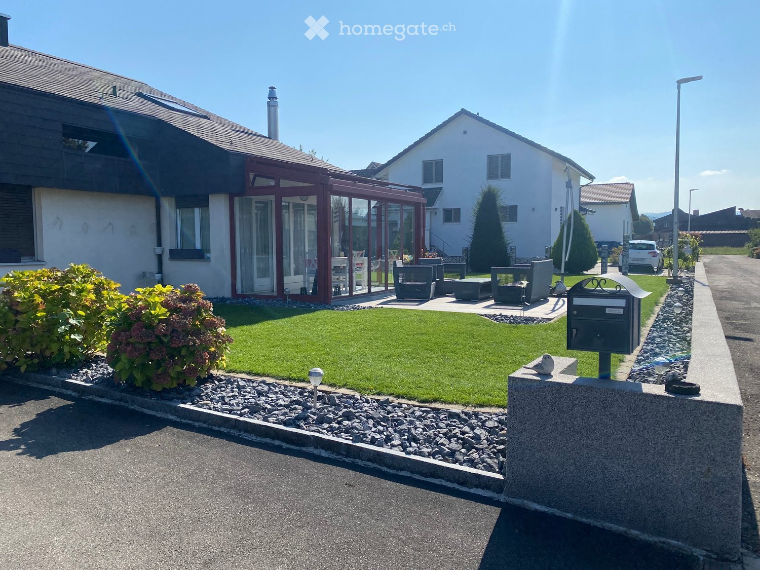 Well-maintained garden with a lawn, patio seating area, and a mailbox. The house features a modern design with large windows and a covered patio area.