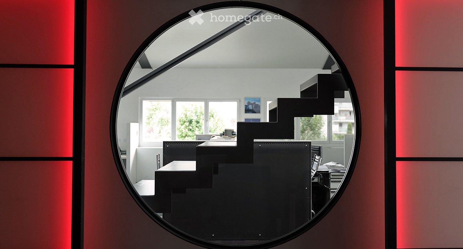 A modern, minimalist room visible through a round mirror, featuring a black chair, a table, a laptop, and a picture frame on the wall.