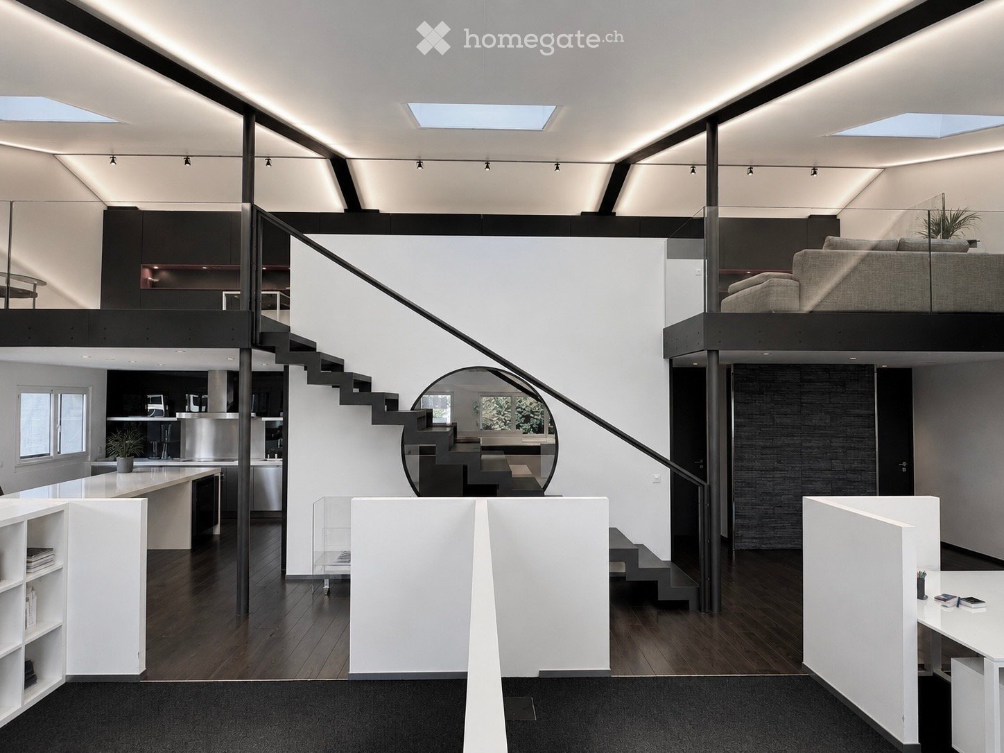 Modern, open-concept living space with a spiral staircase leading to an upper loft area. Features include a sleek kitchen island, a comfortable sofa, a glass-walled staircase, and contemporary furniture.