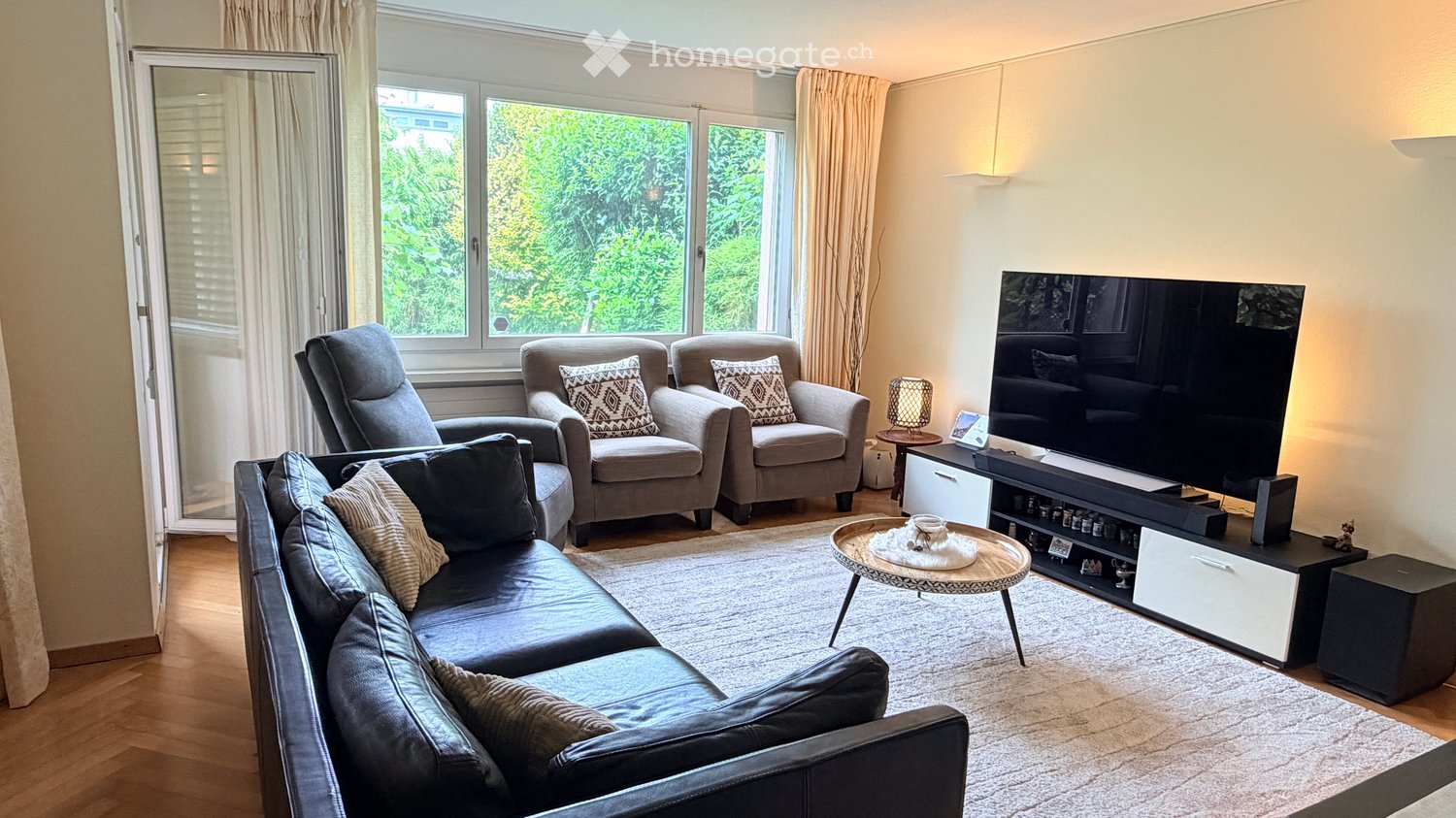 Modern living room with large windows, grey sofa, armchairs, coffee table, TV, and decorative pillows.