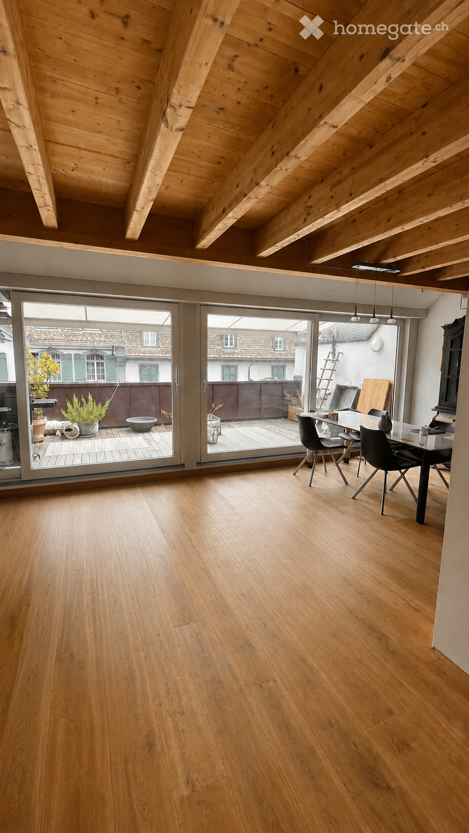Bright, airy room with wooden floors and exposed wooden beams on the ceiling. Large sliding glass doors open to a balcony or terrace area with outdoor furniture.