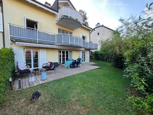 A well-maintained garden area with a wooden deck, outdoor furniture, and a small seating area. The deck is adjacent to a multi-story yellow building with balconies.