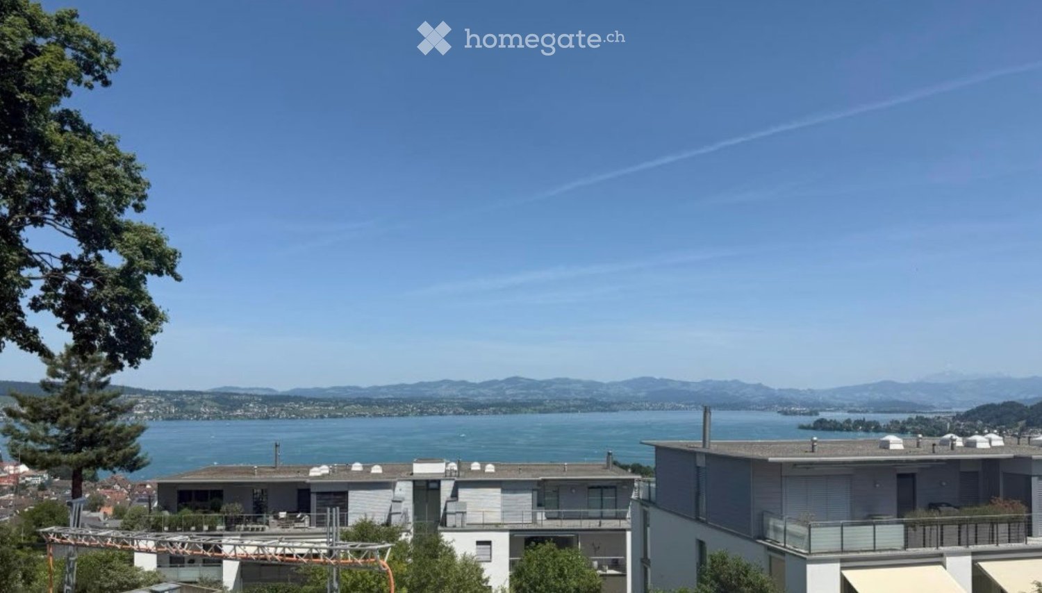 Modern multi-story building with balconies overlooking a scenic lake and distant mountains.