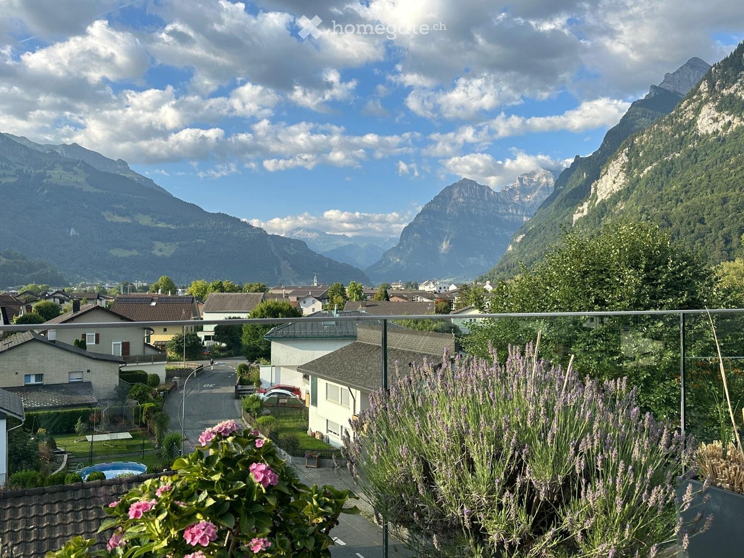 Balcony with a scenic mountain view, surrounded by houses and greenery, with potted flowers in the foreground.