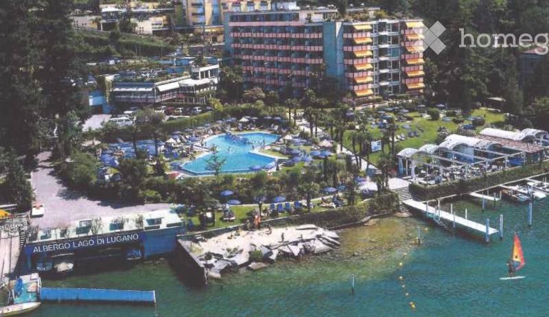 Aerial view of a resort area with a swimming pool, lounge chairs, umbrellas, and a waterfront with boats and a sailboard.