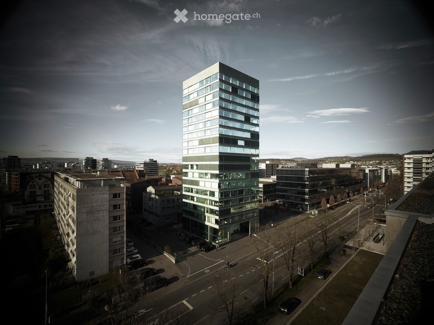 A modern, multi-story building with large glass windows and a sleek design, surrounded by other structures and a parking area.