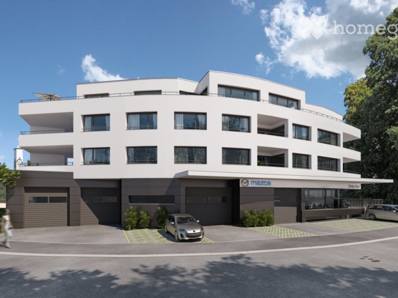 Modern two-story building with white facade, large windows, and multiple balconies. Features a covered parking area with several cars and a Mazda logo visible on the building.