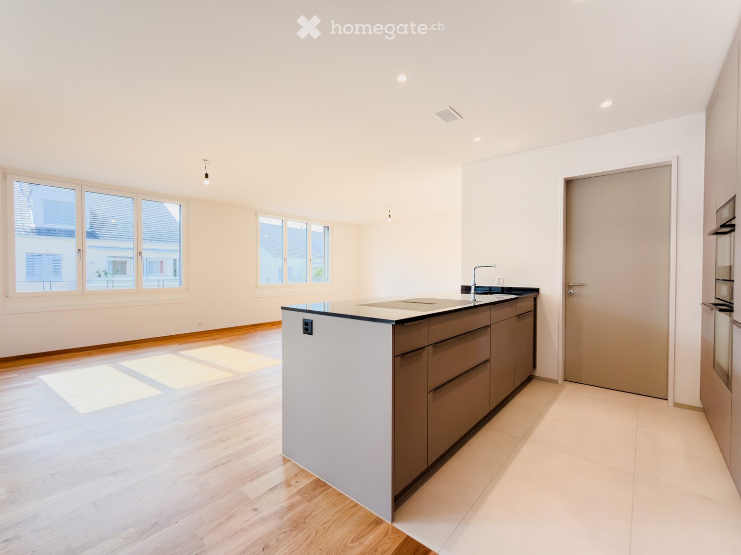 Empty kitchen with modern design, featuring a central island, wooden flooring, and large windows allowing natural light to fill the space.