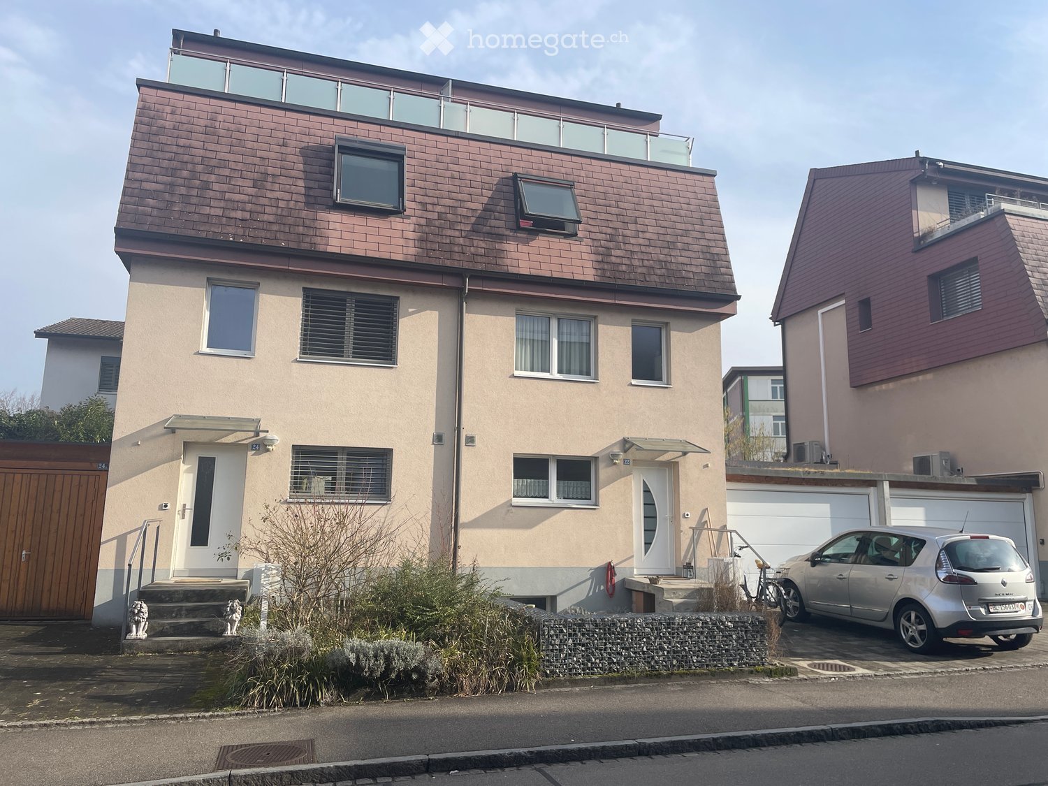 Modern two-story house with a brown tiled roof, large windows, and a balcony on the upper level. The house has a light beige facade and a small garden area in front.