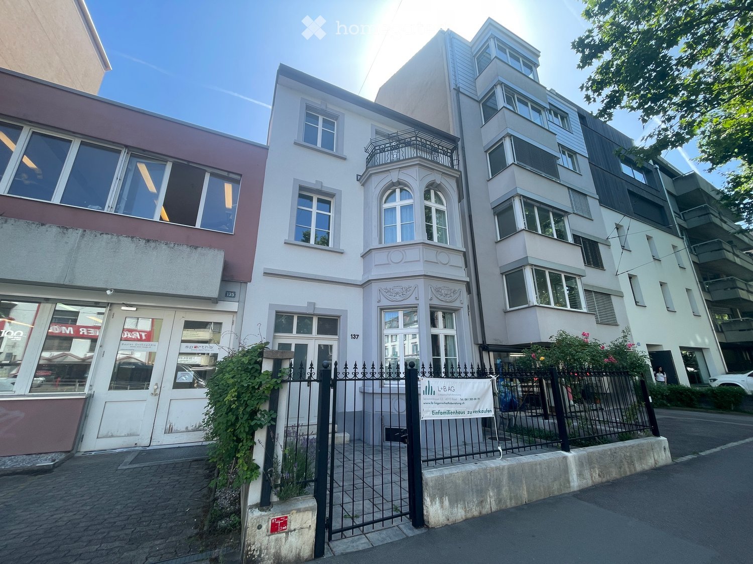 Multi-story residential building with white facade, arched windows, and decorative balcony. The entrance features a black wrought-iron fence and signage for a real estate agency.